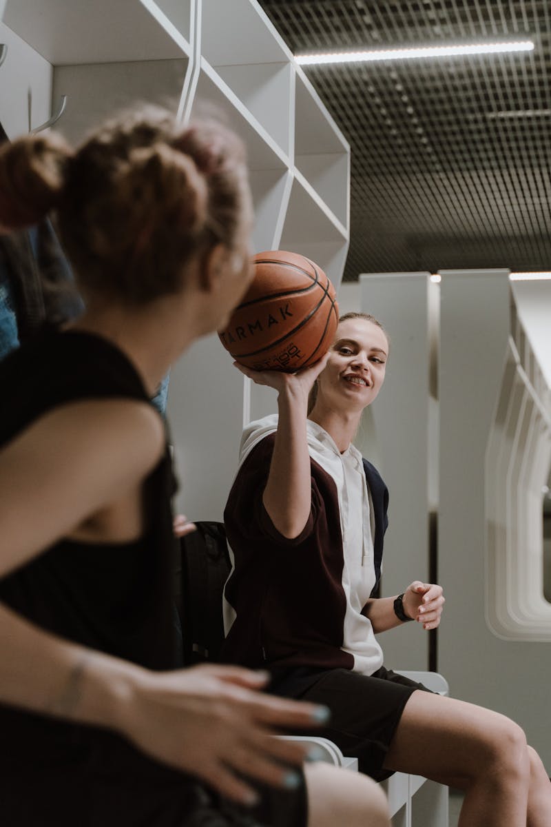 Two young women enjoying a playful moment with a basketball in a locker room setting.
